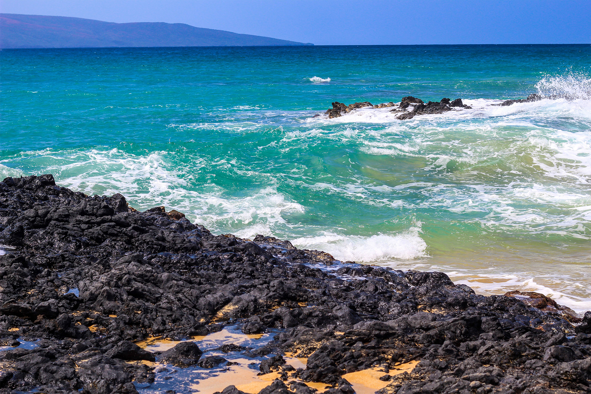 A beach with waves crashing on it and rocks in the foreground.